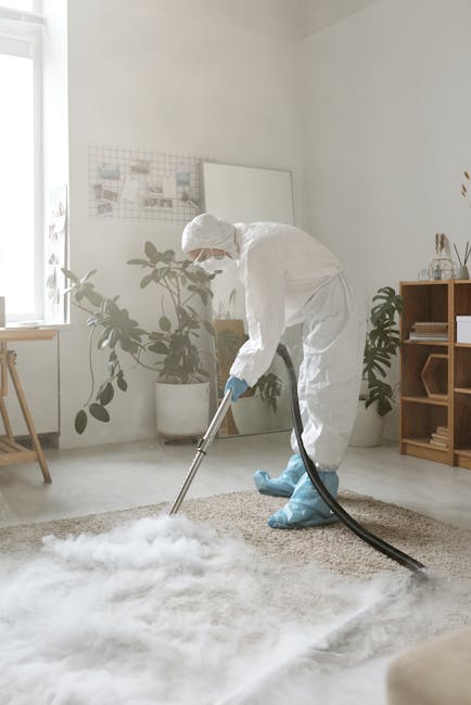 A professional cleaner dressed in a white protective suit, face mask, and blue gloves is performing deep cleaning on a beige carpet in a bright, modern living room. The cleaner is using a steam or foa