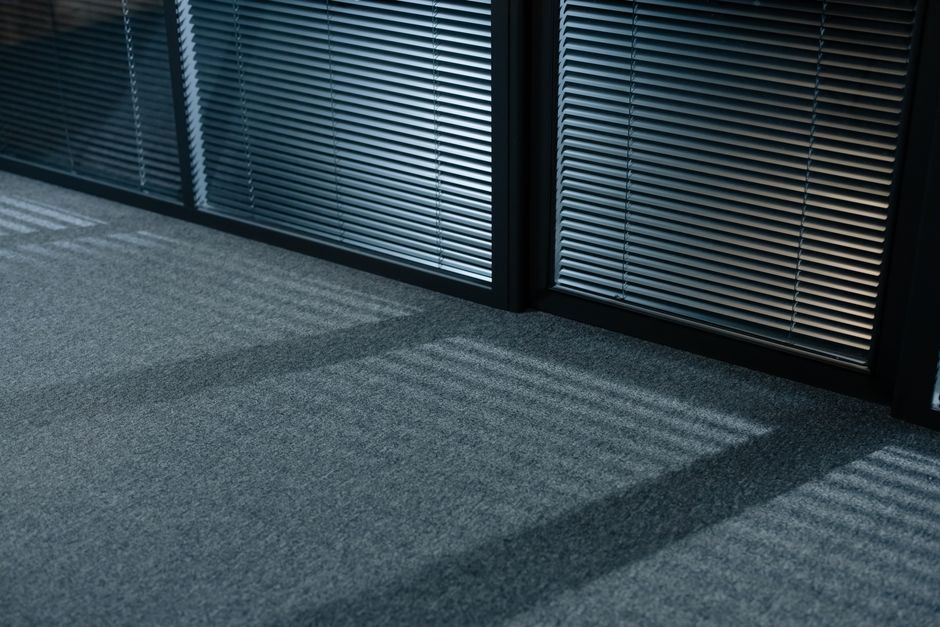 A clean and well-lit corridor in a commercial building featuring a grey carpeted floor with a subtle textured pattern, white walls with several doorways, and a white ceiling with evenly spaced recesse