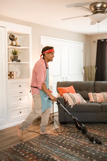 A person vacuuming a living room carpet with a handheld vacuum cleaner, wearing casual clothing and an apron, in a well-lit space with neutral-colored walls, a gray sofa with orange and patterned cushions, a patterned area rug, a white shelving unit with decorative items including books and plants, a ceiling fan, and a window with dark curtains, showcasing surface cleaning and maintenance typical of domestic cleaning services by Merton Park Carpet Cleaners.