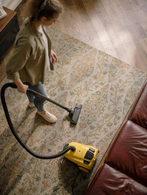 A person vacuuming a living room carpet with a handheld vacuum cleaner, wearing casual clothing and an apron, in a well-lit space with neutral-colored walls, a gray sofa with orange and patterned cushions, a patterned area rug, a white shelving unit with decorative items including books and plants, a ceiling fan, and a window with dark curtains, showcasing surface cleaning and maintenance typical of domestic cleaning services by Merton Park Carpet Cleaners.