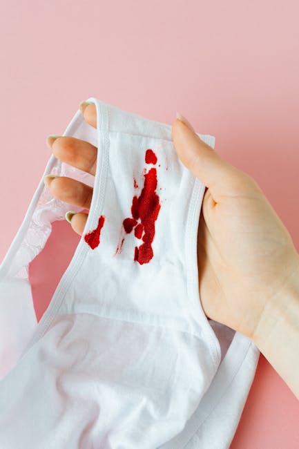 A person holding a white fabric mask with visible red bloodstains on a pink background, demonstrating cleaning or stain removal processes. This image emphasizes the importance of surface cleaning and sanitation, relevant to domestic and commercial cleaning services offered by Merton Park Carpet Cleaners, as highlighted in the webpage 'Urgent spill? Same-day stain removal in Merton Park'. The hand is gripping the mask firmly, with a focus on the stained areas, and appears to be in a well-lit environment, suggesting a clean and hygienic setting suitable for professional stain and spill removal.